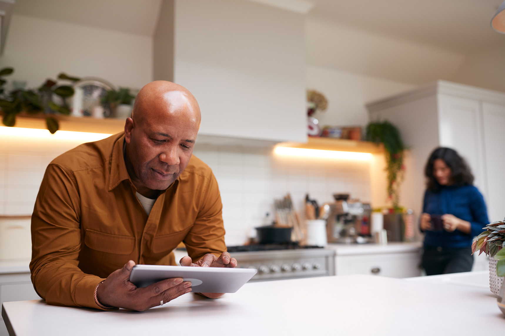 A mature man seated at a kitchen island staring at a digital tablet and a woman standing in the background looking at her phone