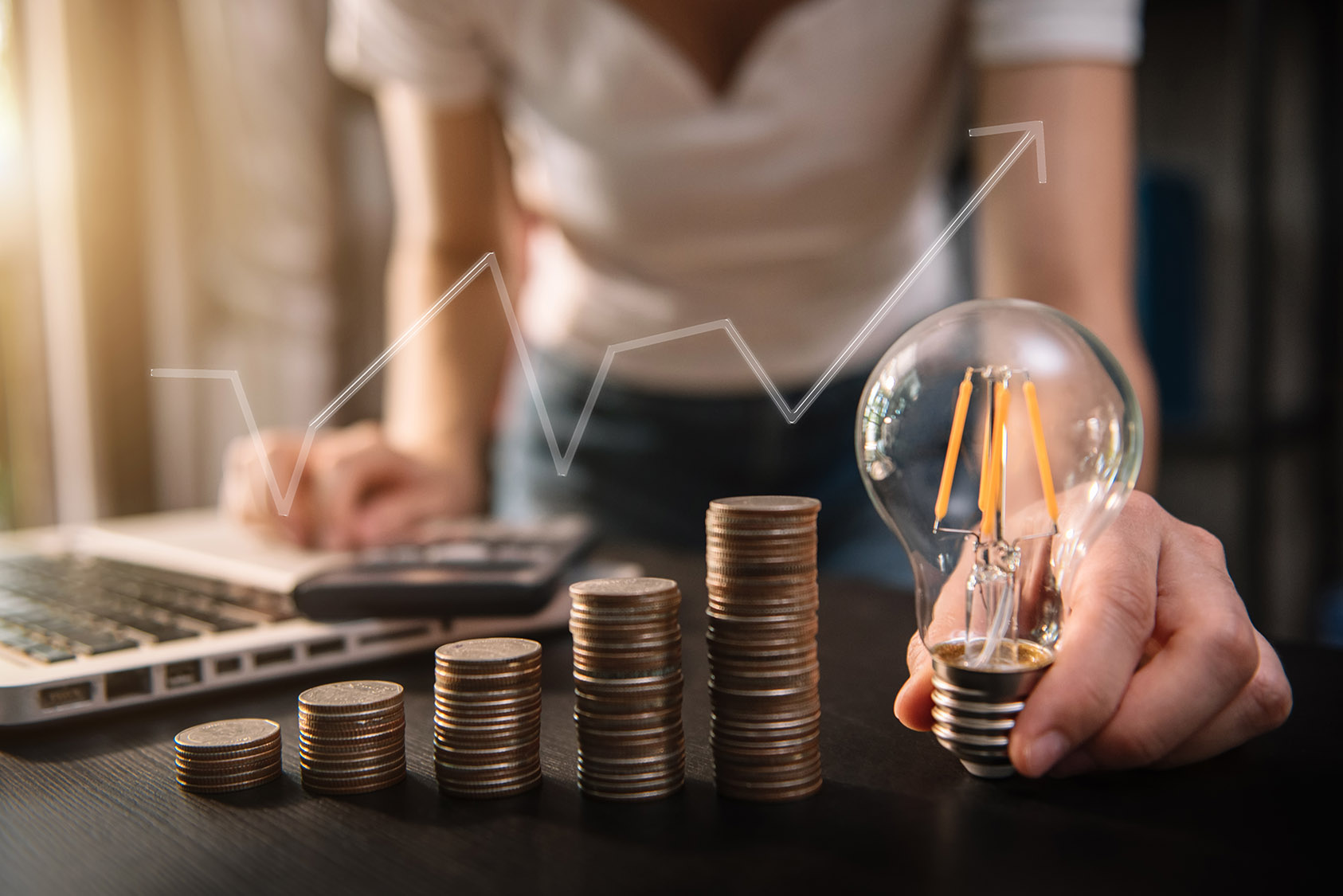 Photo of a person holding an LED lightbulb with columns of coins stacked and a laptop and calculator in the background