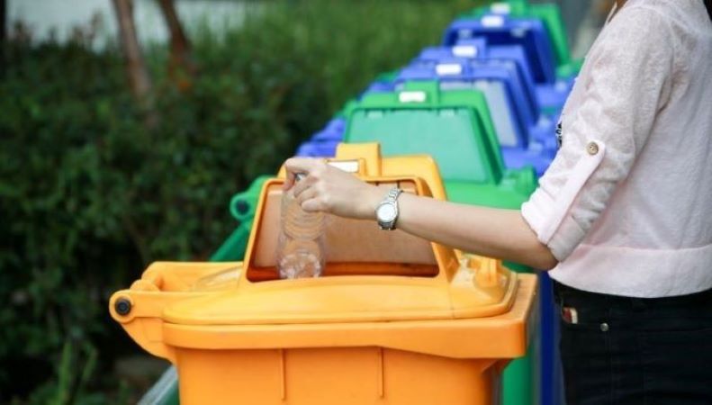 Person placing a clear drinking container in a recycle bin near various colored recycle bins.